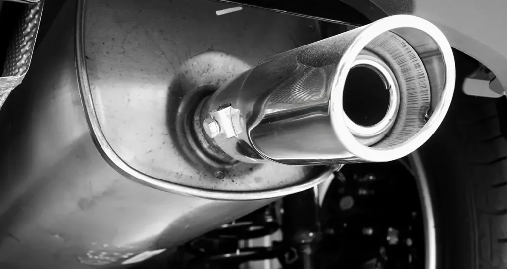 Nahaufnahme eines verchromten Auspuffendrohrs am Schalldämpfer eines Pkw; Unterboden und Radpartie im Hintergrund.
