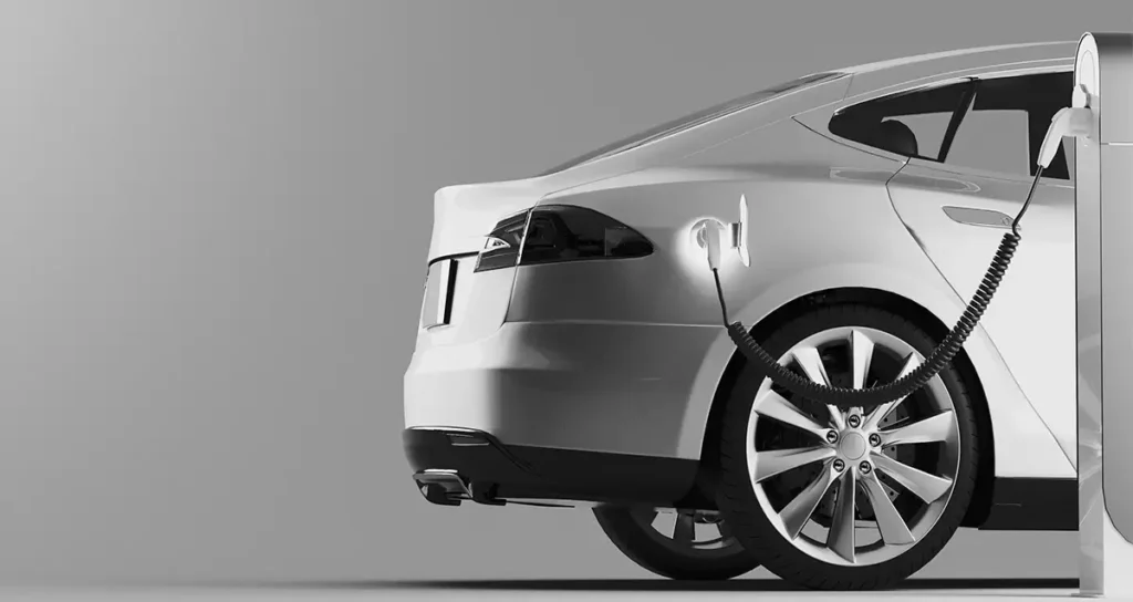 Teilansicht eines weißen Elektroautos im Studio: Heck und rechtes Hinterrad, Ladekabel an einer Säulen-Ladestation eingesteckt vor grauem Hintergrund.