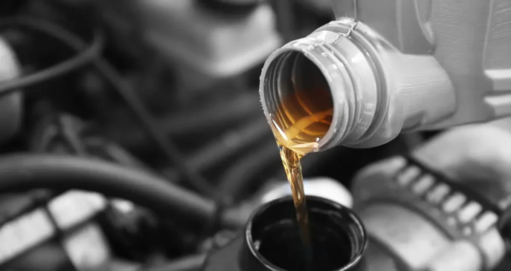 Nahaufnahme: goldfarbenes Motoröl fließt aus einem Kunststoffkanister in die Einfüllöffnung eines Motors; unscharfer Maschinenraum im Hintergrund.