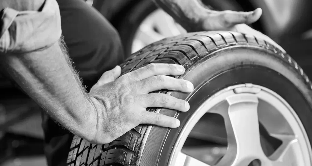 Nahaufnahme: Mechaniker prüft mit beiden Händen Profil und Sitz eines Pkw-Reifens auf der Felge; Reifenservice in Schwarz-Weiß.