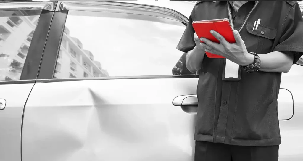 Gutachter/Mechaniker dokumentiert mit rotem Tablet eine deutliche Delle in der Autotür eines silbernen Fahrzeugs.